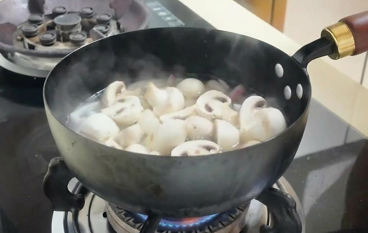 Sliced white mushrooms and cubes of tofu simmering in a bubbling broth inside a dark cooking pan on a stove.