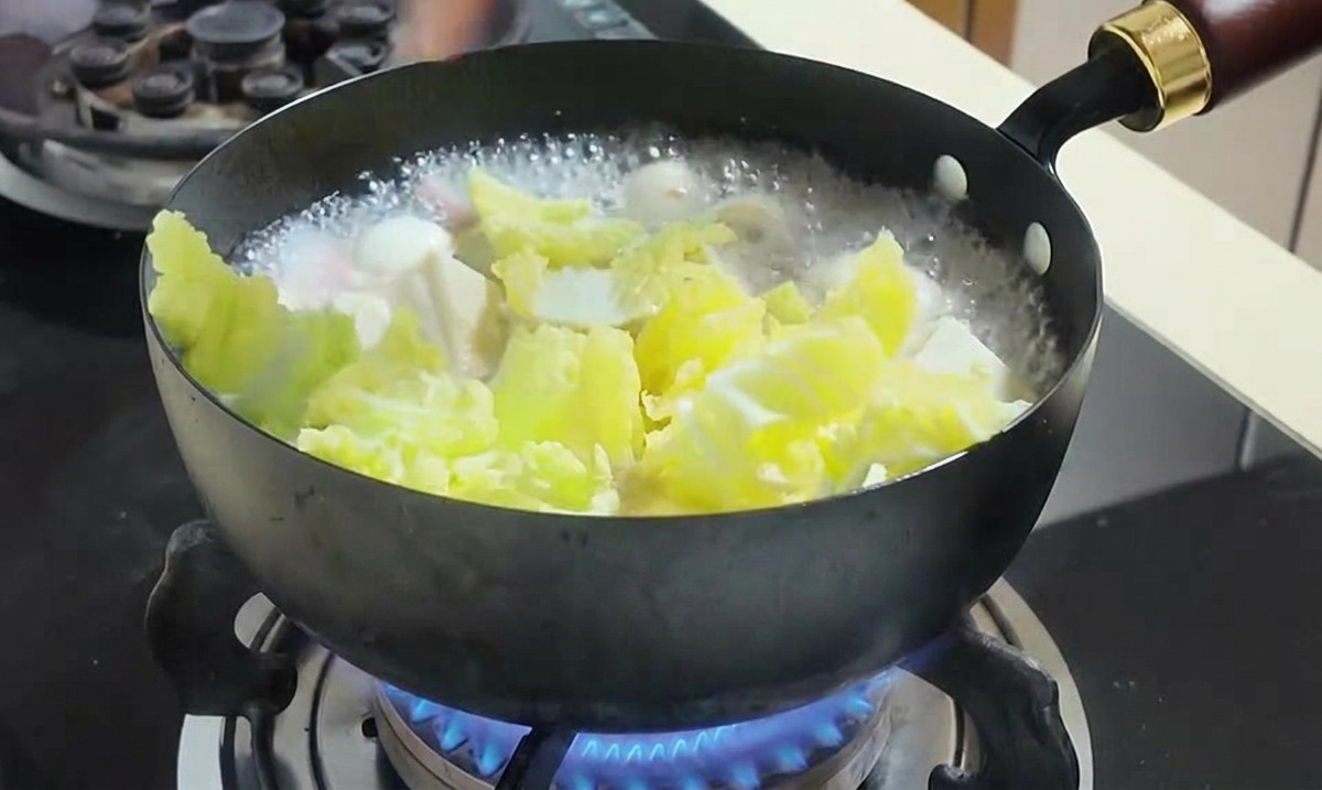 A hand adding bright yellow corn kernels and chopped green cabbage leaves into a boiling pot of soup.