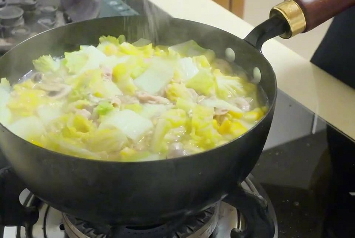 A hand sprinkling fine white salt into a steaming pan filled with vegetables, tofu, and tuna soup.