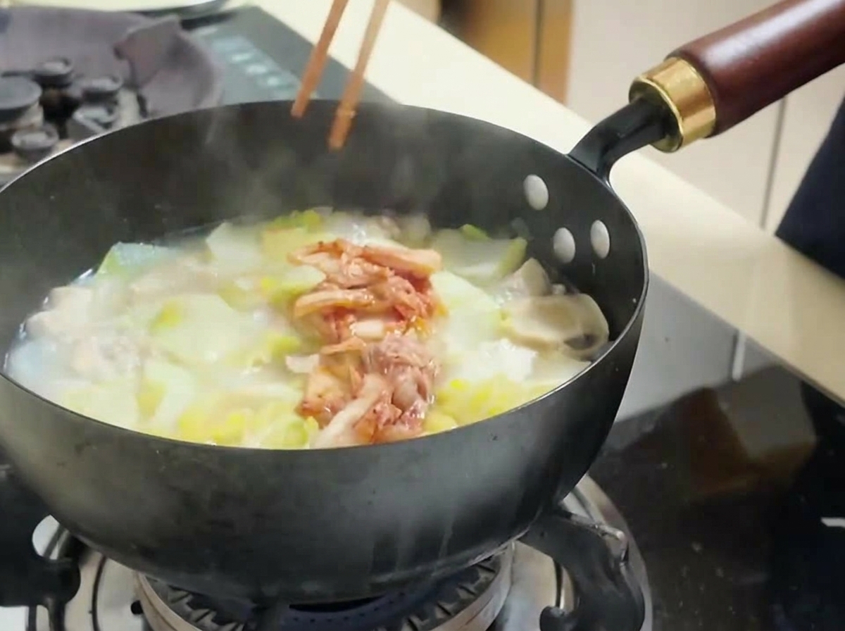 A person using chopsticks to add a portion of red chopped kimchi into a simmering pot of soup with tofu and cabbage.