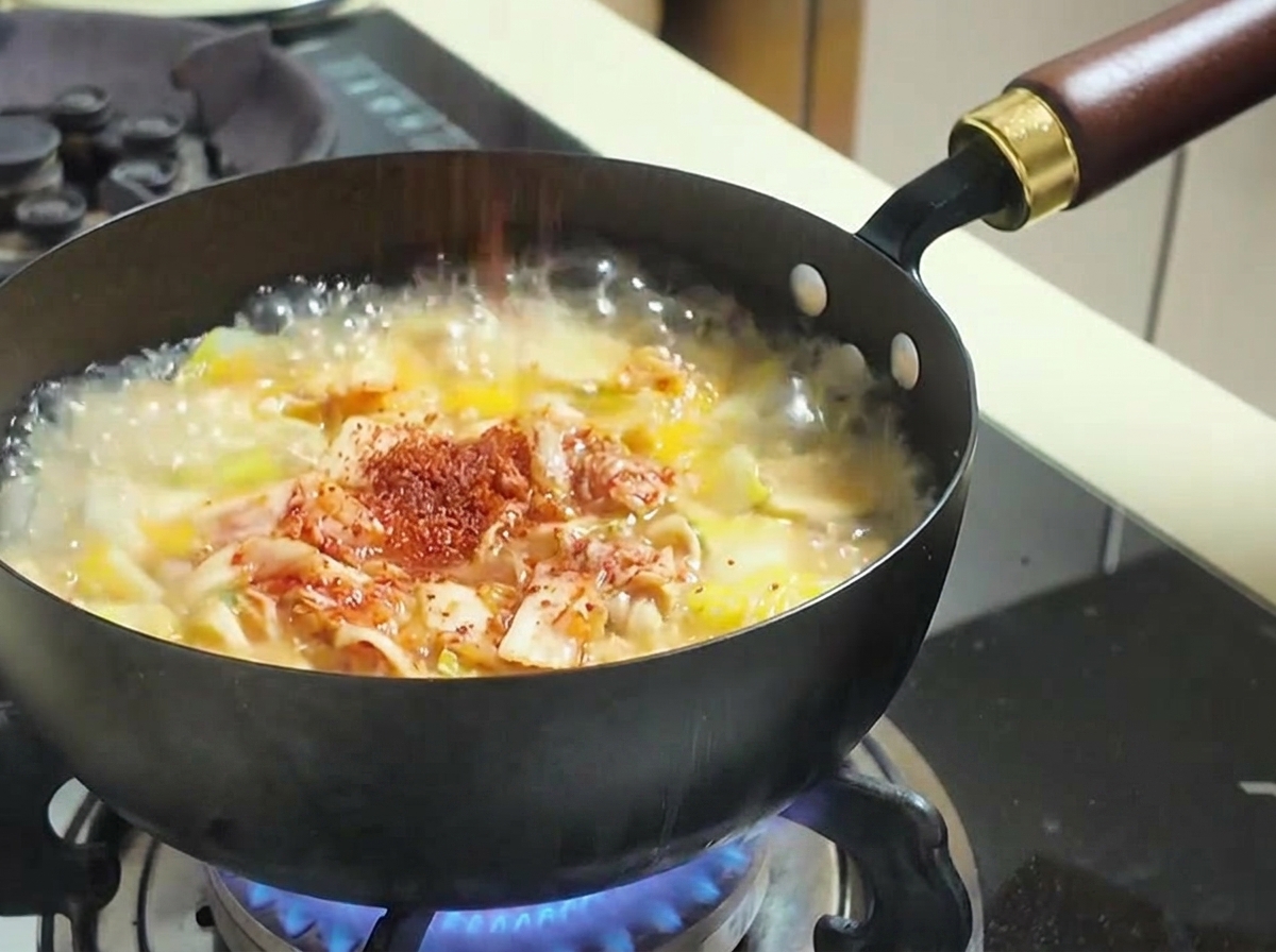 Red chili powder being sprinkled into a bubbling pot of soup containing kimchi and vegetables.
