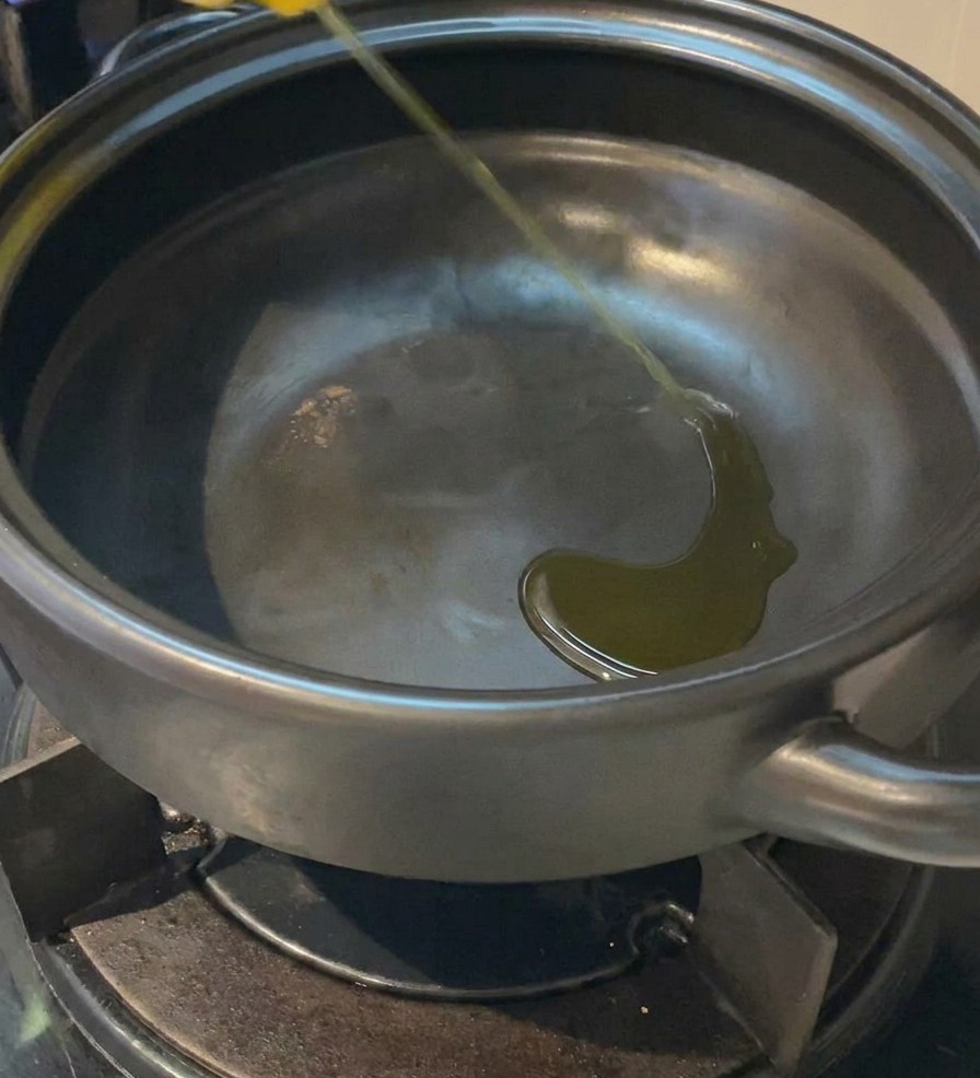 A thin stream of yellow cooking oil being poured into the bottom of a dark, empty clay pot sitting on a gas stove.