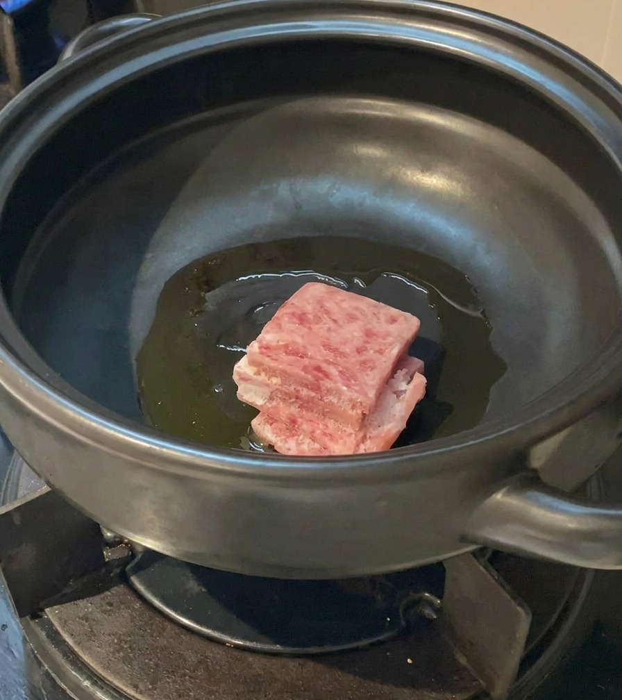 Slices of raw, marbled pork belly being placed into a clay pot with shimmering oil.