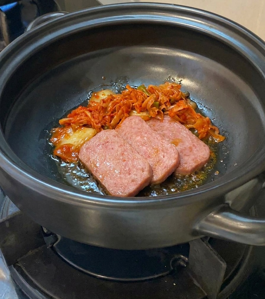 Chopped kimchi being added to a clay pot containing partially cooked slices of meat.