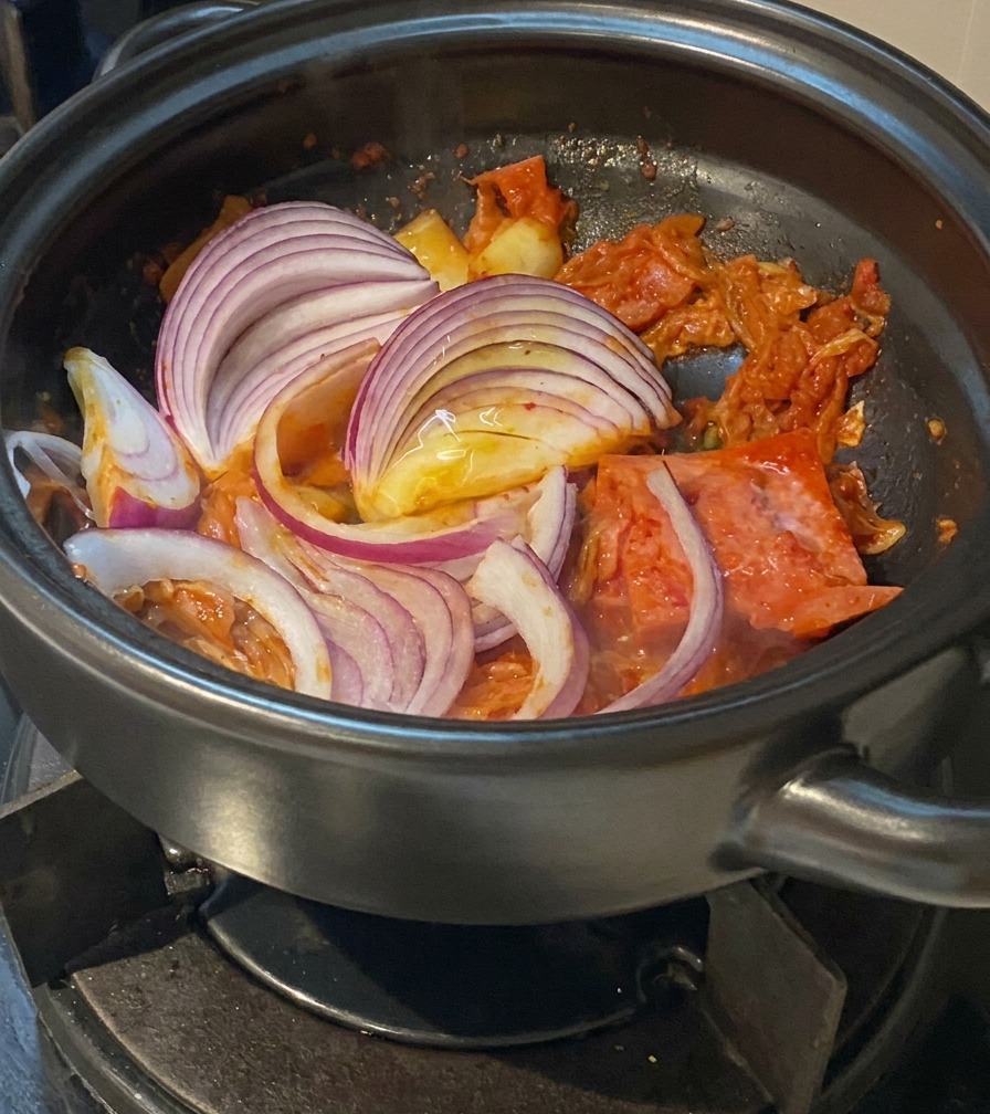 Thick crescents of purple red onion being stirred into a pot of sautéed kimchi and pork.