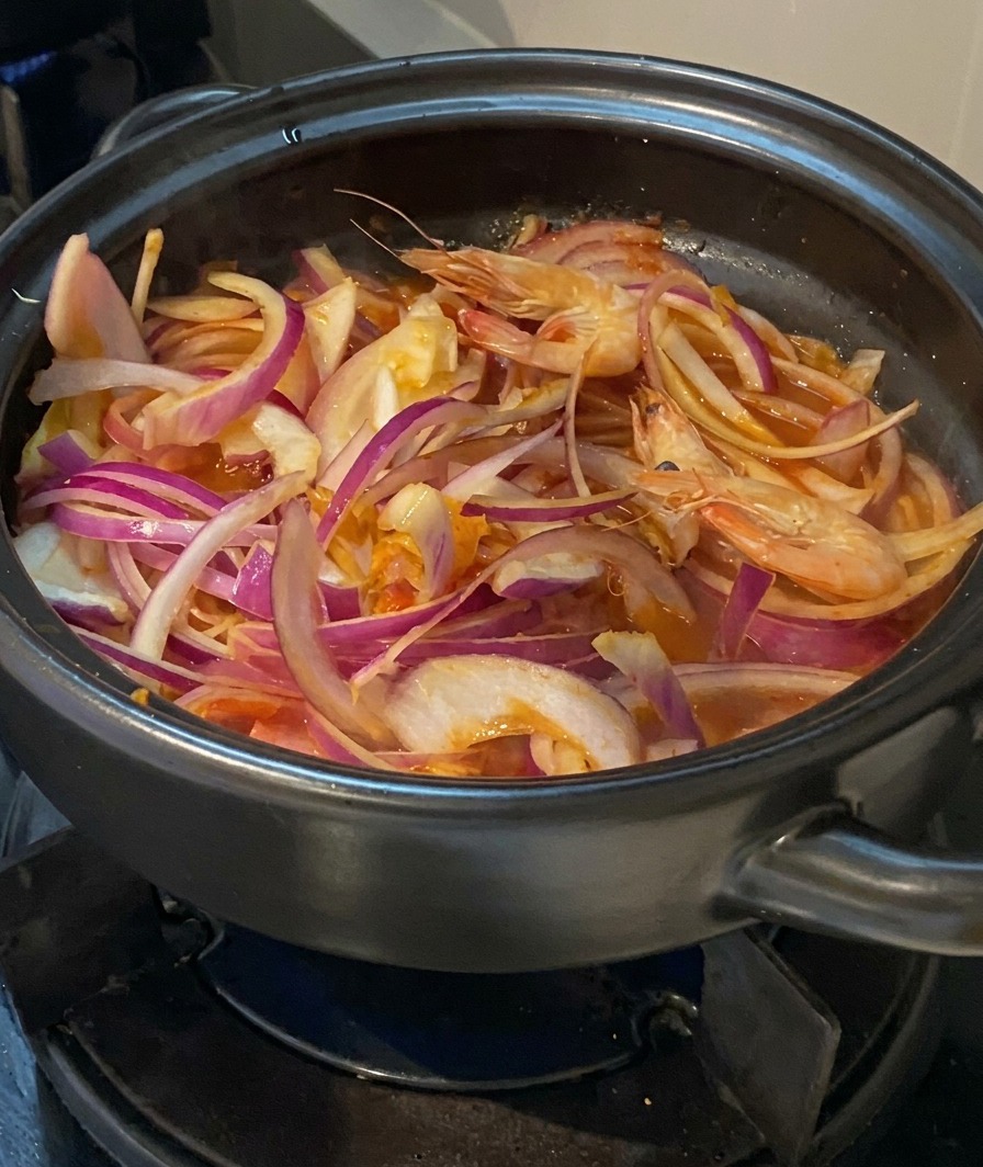 Fresh whole shrimp with heads on being added to a pot filled with onions, kimchi, and pork.