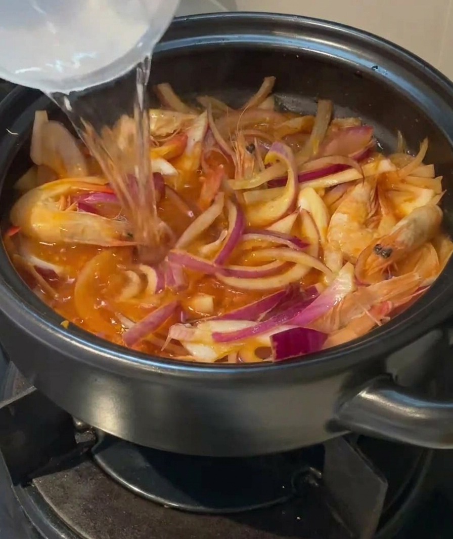 Clear water being poured from a container into a clay pot filled with shrimp, onions, and kimchi.