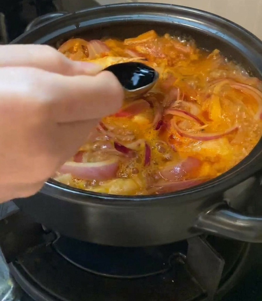 A hand holding a metal spoon and pouring dark light soy sauce into a bubbling pot of kimchi soup with sliced red onions.