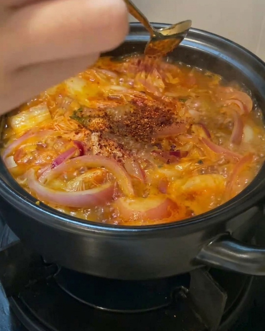A hand using a gold spoon to sprinkle bright red chili powder over a bubbling pot of kimchi and onion soup.