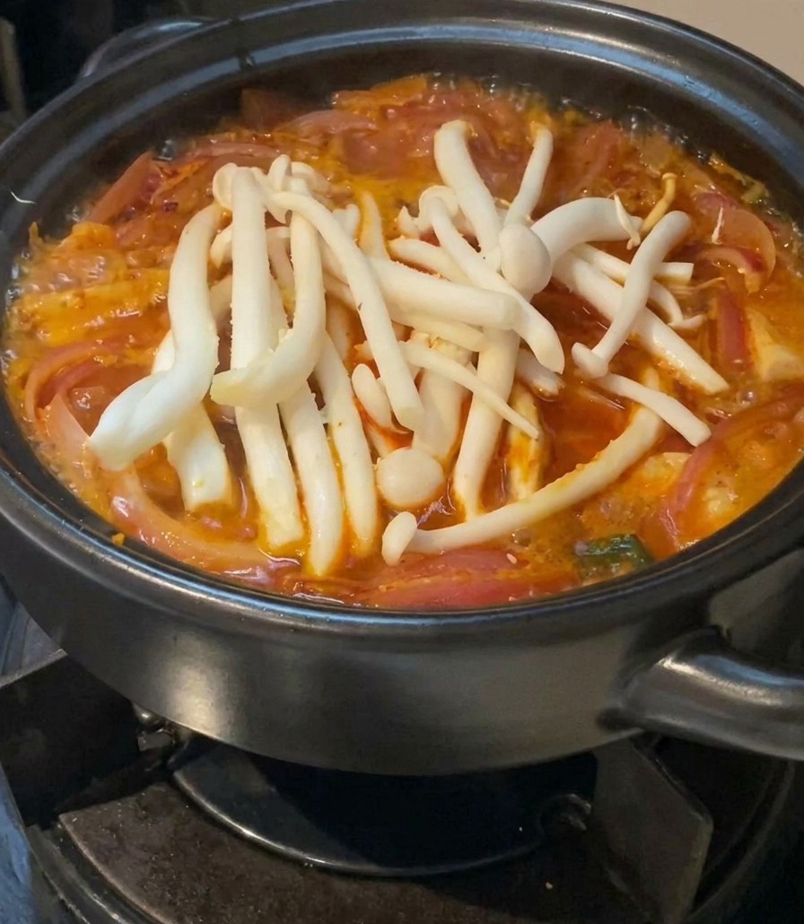 A cluster of fresh, white seafood mushrooms resting on top of a red, simmering kimchi soup in a black ceramic pot.