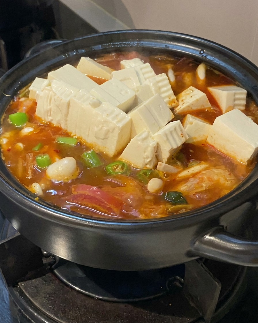 Numerous cubes of fresh white tofu and sliced green chili peppers being added to a reddish orange bubbling soup in a black ceramic pot.