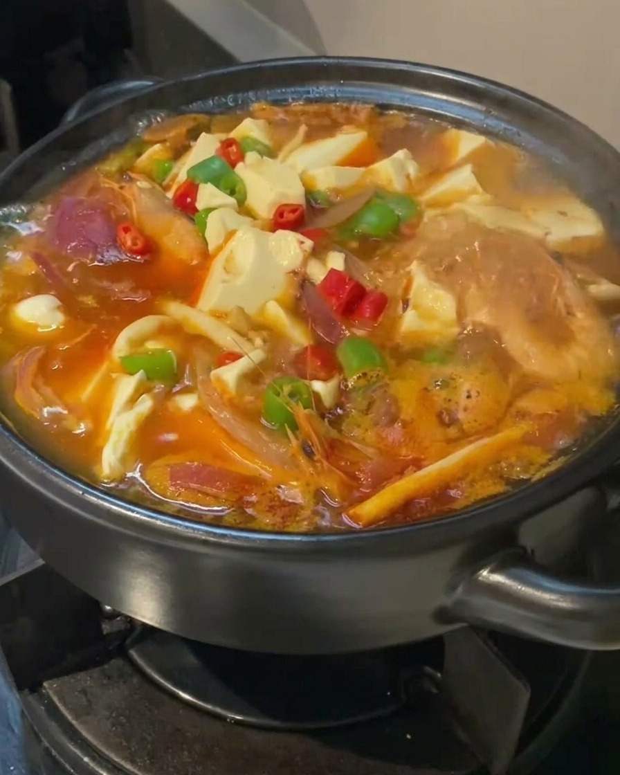 A close-up of Kimchi Tofu soup simmering in a black pot, garnished with bright red and green sliced chilies on top of white tofu cubes.