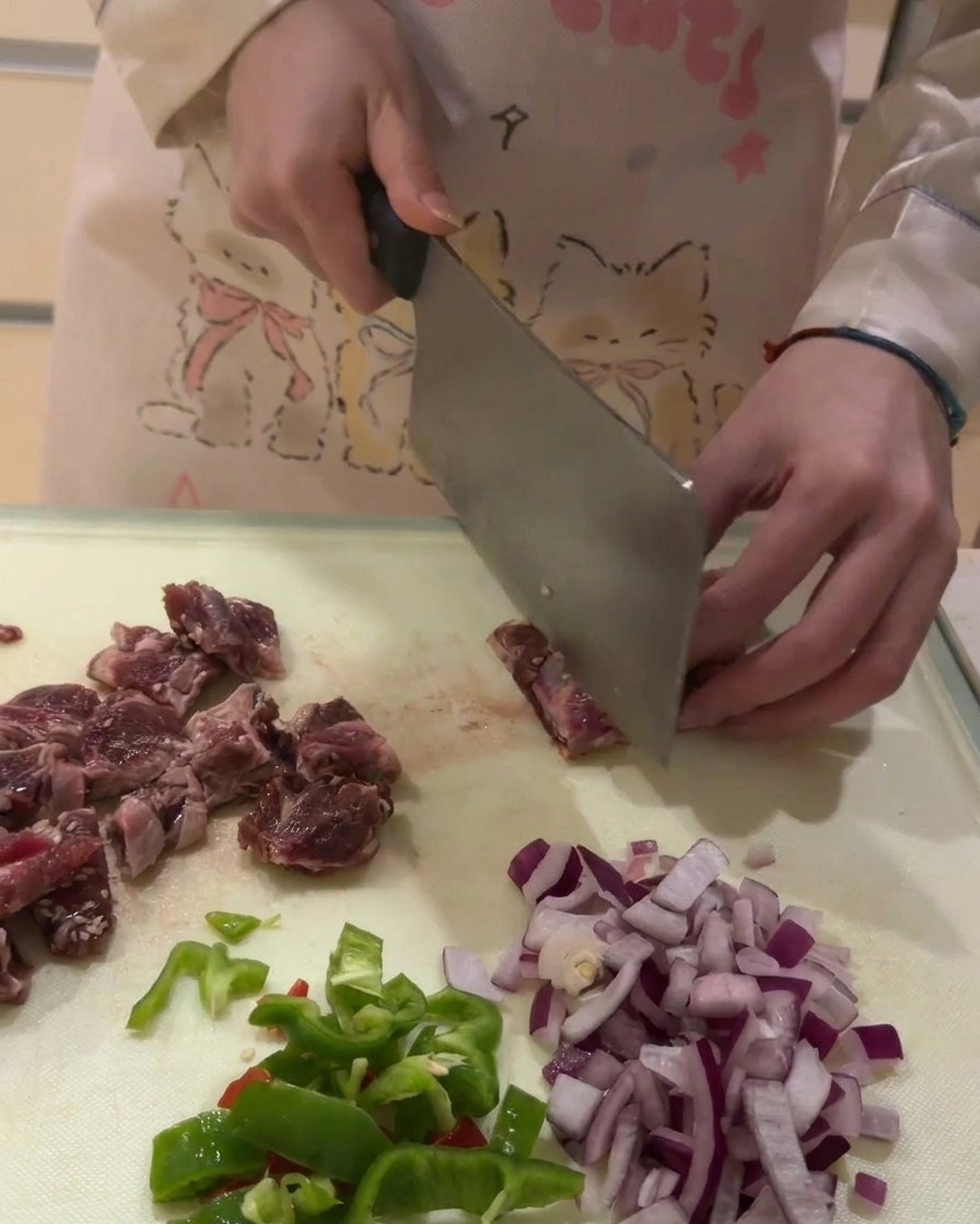 A close-up shot of raw beef rib meat being sliced into small square chunks on a white cutting board.