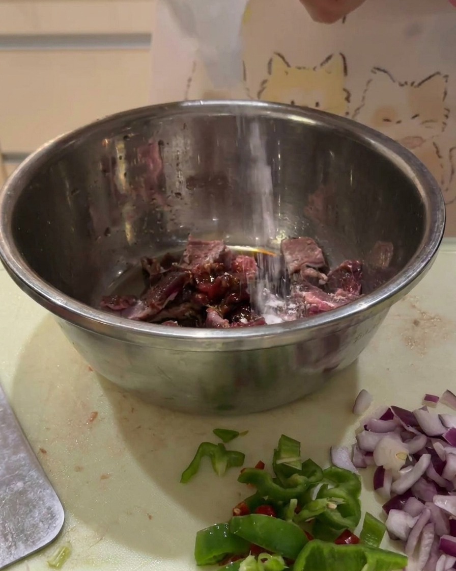 Dark sauces being poured from a bottle onto raw beef cubes in a metal mixing bowl.