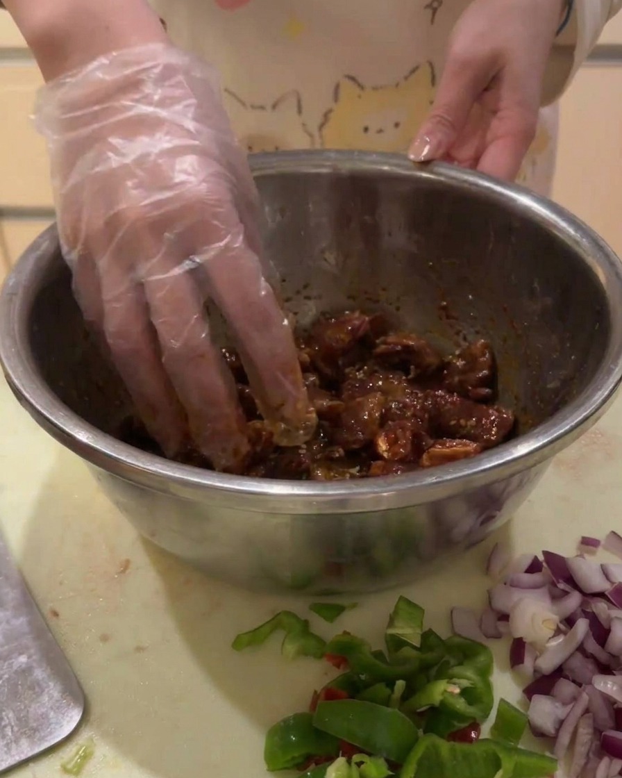 A gloved hand mixing marinated beef cubes in a silver bowl to ensure even coating.