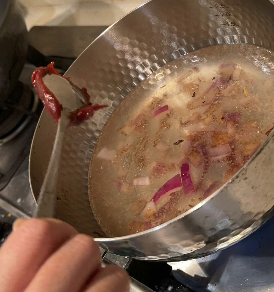 A metal spoon lowering a large dollop of thick red chili paste into the water-filled pot with onions.