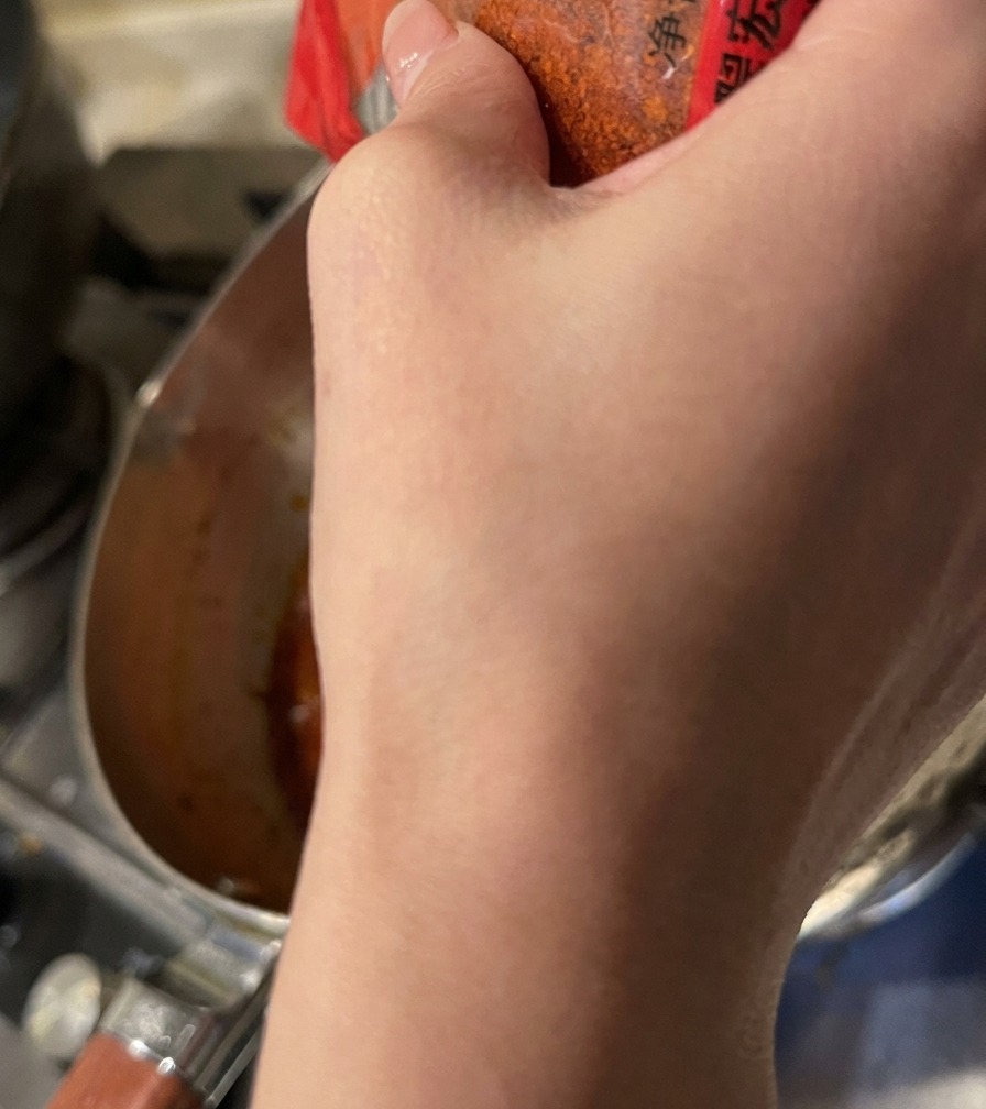 Red chili powder being sprinkled from a container into the simmering orange soup base.