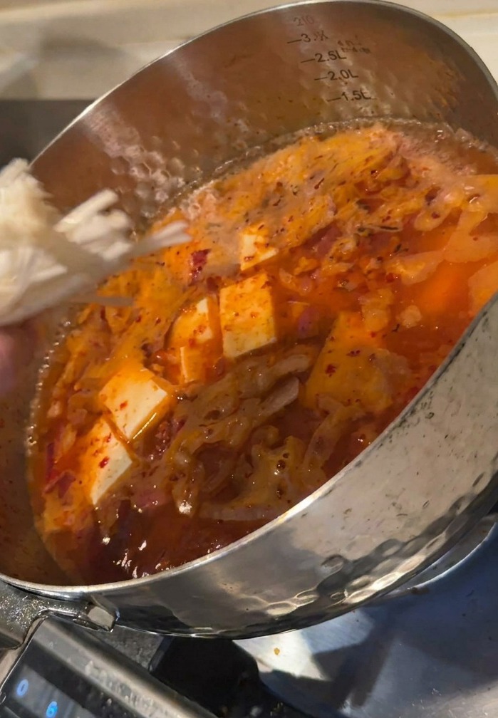 A hand adding a bundle of white enoki mushrooms into a pot of bubbling red kimchi soup containing cubes of tofu.