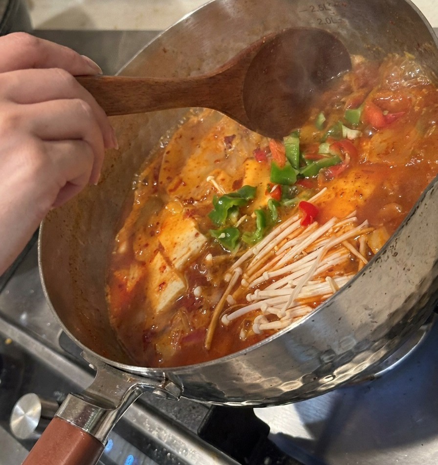 Sliced green peppers and fermented kimchi being added to the bubbling soup and stirred with a wooden spoon.