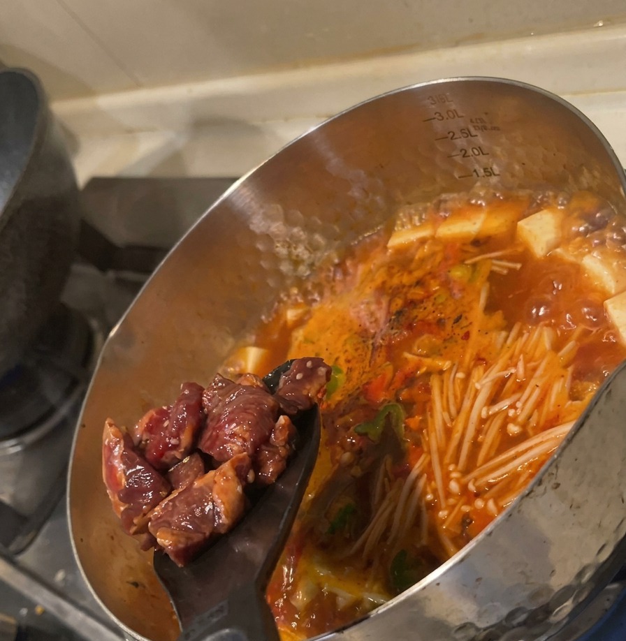Raw marinated beef cubes being pushed from a black spatula into a boiling red soup filled with vegetables.