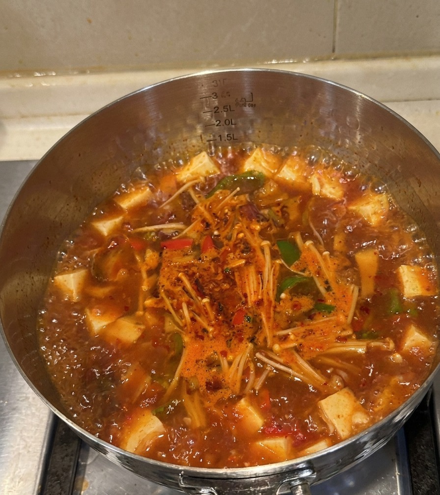 A stainless steel pot filled with bubbling red kimchi beef soup, showing cooked meat, enoki mushrooms, and peppers.
