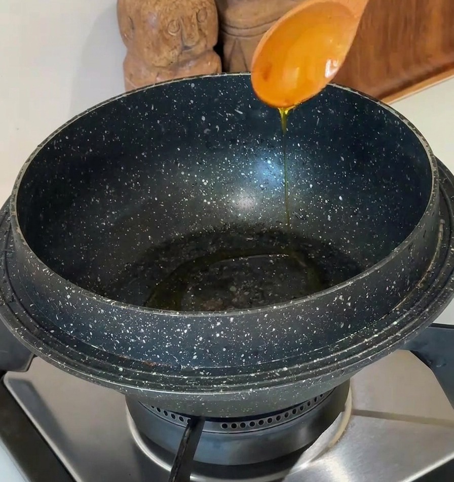 Cooking oil being poured from a light-colored wooden spoon into a dark, speckled stone pot on a modern stovetop.