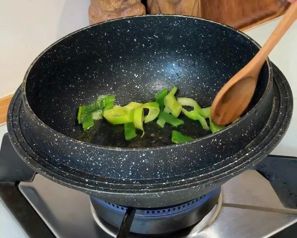 Chopped green scallions being stirred with a wooden spoon in a dark, granite-patterned pot on a gas stove.