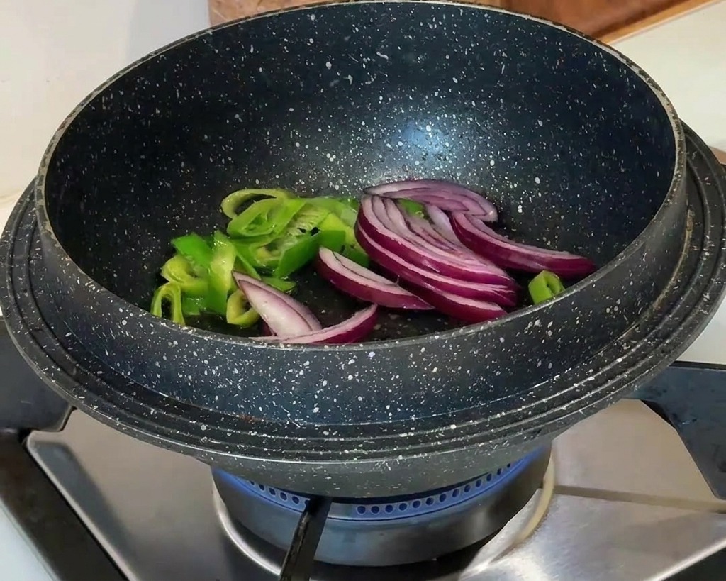 Slices of red onion being added to sautéed green scallions in a black pot over a blue gas flame.