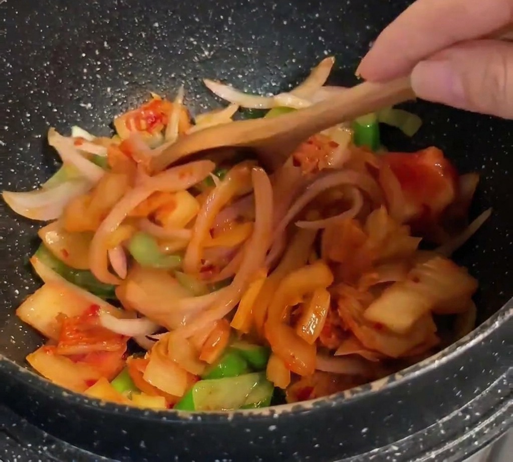 A wooden spoon stirring bright orange-red kimchi into a mixture of sautéed onions and scallions.