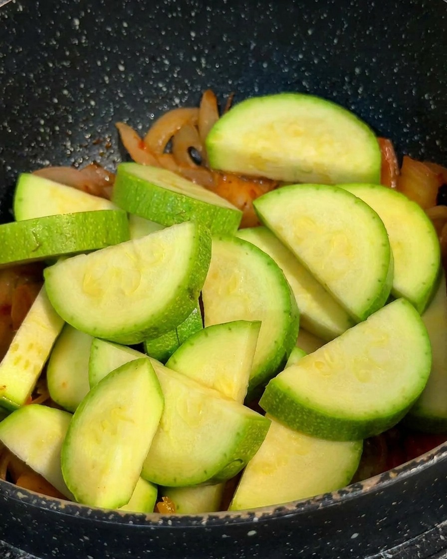 Thick half-moon slices of fresh green zucchini piled into a pot over a base of cooked kimchi and onions.