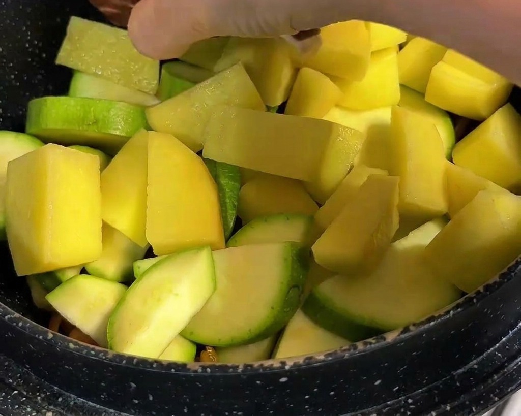 Chunks of yellow potato and whole shiitake mushrooms with cross-cut patterns being added to a pot of vegetables.