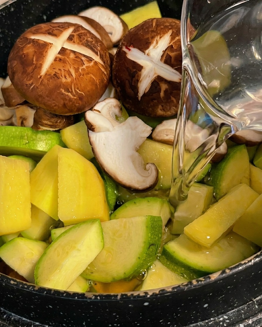 Clear water being poured from a pitcher onto a colorful mix of sliced zucchini, potatoes, and scored shiitake mushrooms in a cooking pot.