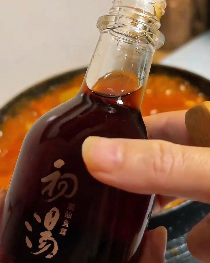A dark glass bottle pouring savory fish sauce into a pot of bubbling red tofu and vegetable stew.