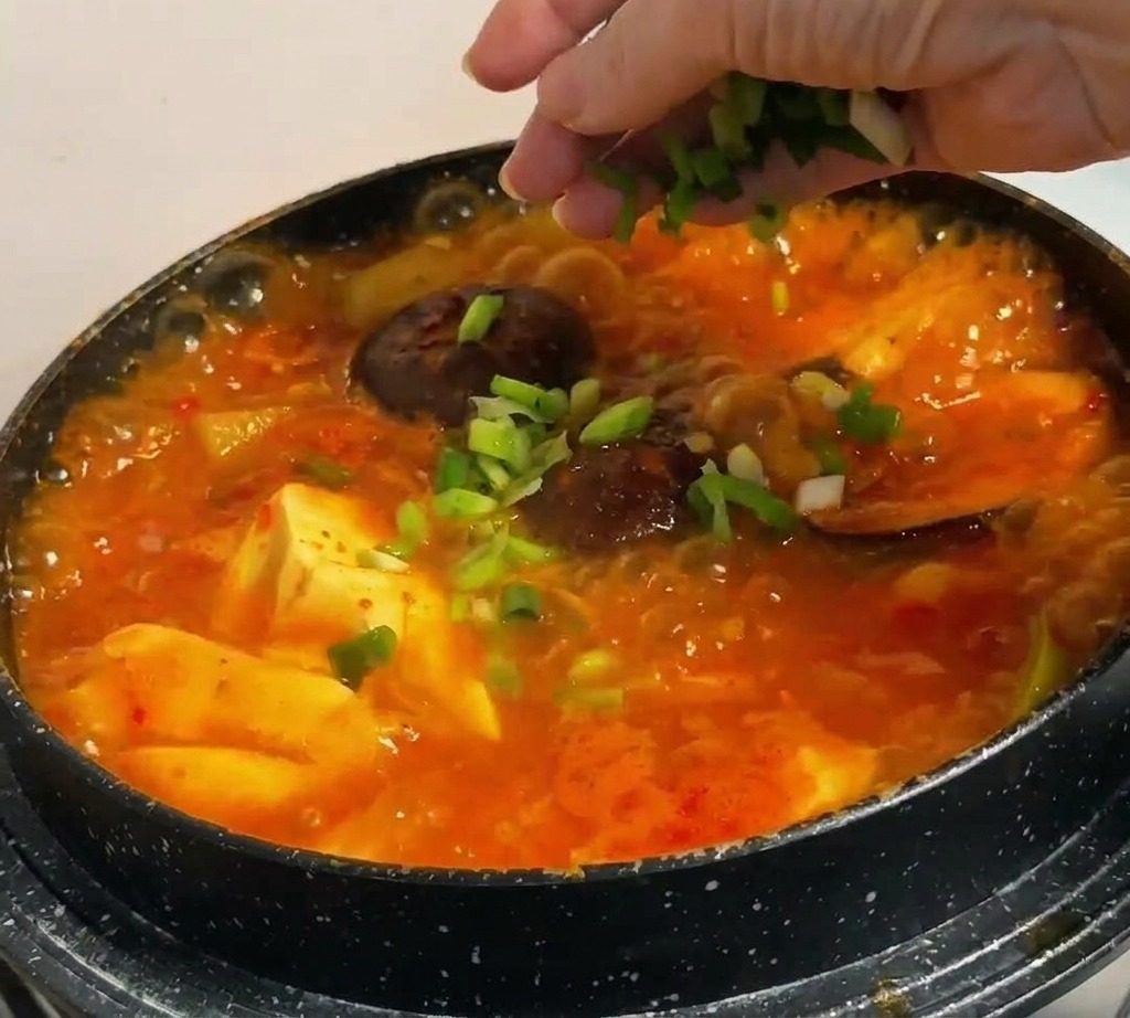 Chopped green onions being sprinkled by hand over a bubbling, vibrant red Korean stew containing tofu and mushrooms.