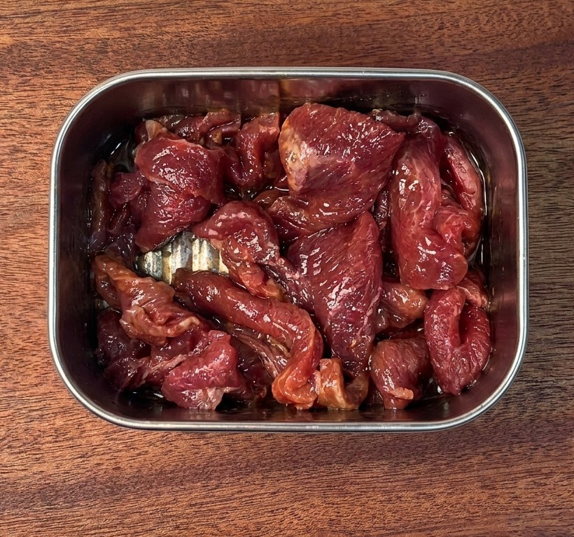 Marinated raw beef slices coated in a glossy dark sauce inside a stainless steel container on a wooden board.