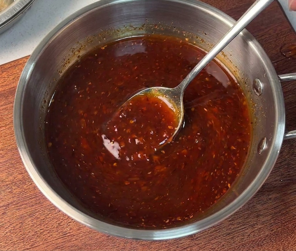 A person stirring a vibrant, thick red sauce base in a small stainless steel bowl with a metal spoon.