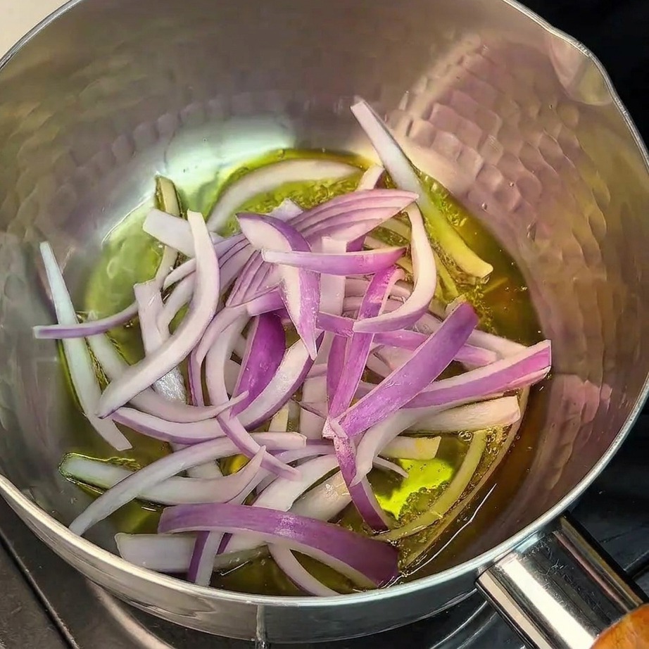 Thinly sliced red onions being stir-fried in oil at the bottom of a hammered stainless steel pot.