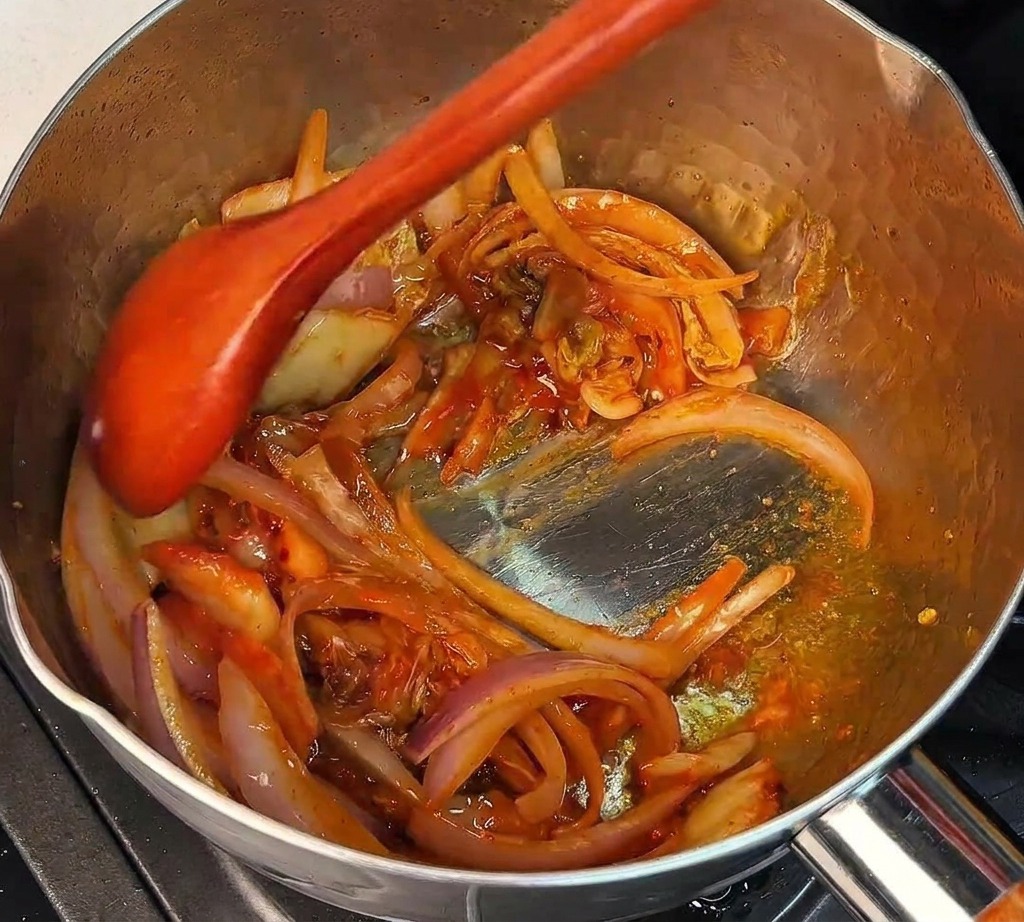 A wooden spoon stirring chopped kimchi and red onions together in a hot metal pot.