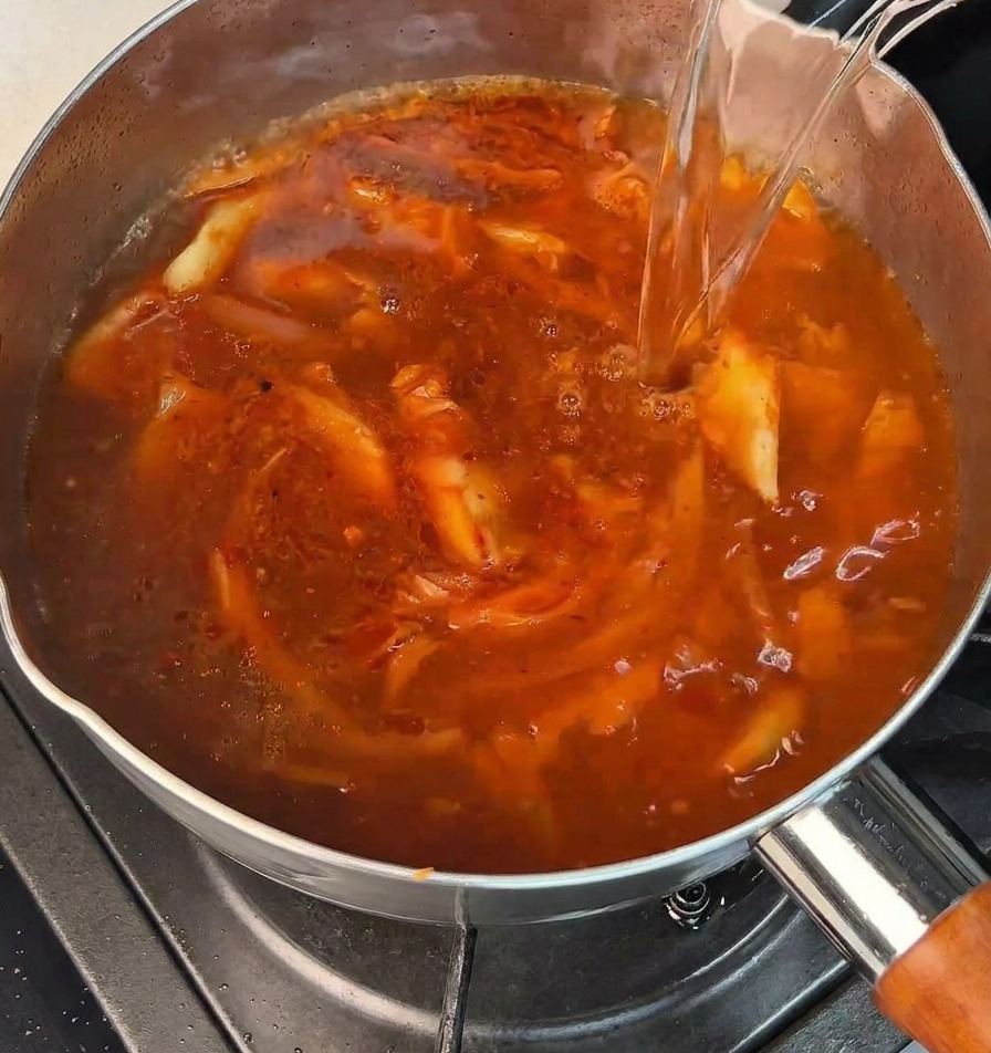 A stream of clear boiling water being poured into a pot with a bubbling red spicy base.