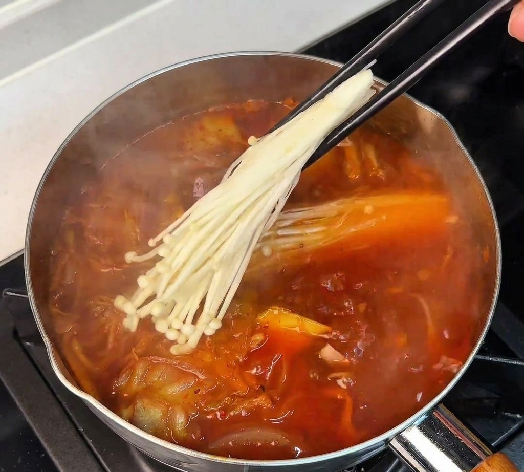 A hand using chopsticks to lower a bundle of white enoki mushrooms into a simmering pot of red kimchi soup.