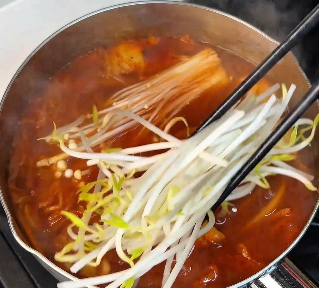 Fresh white bean sprouts being added with chopsticks into the boiling red soup pot alongside enoki mushrooms.