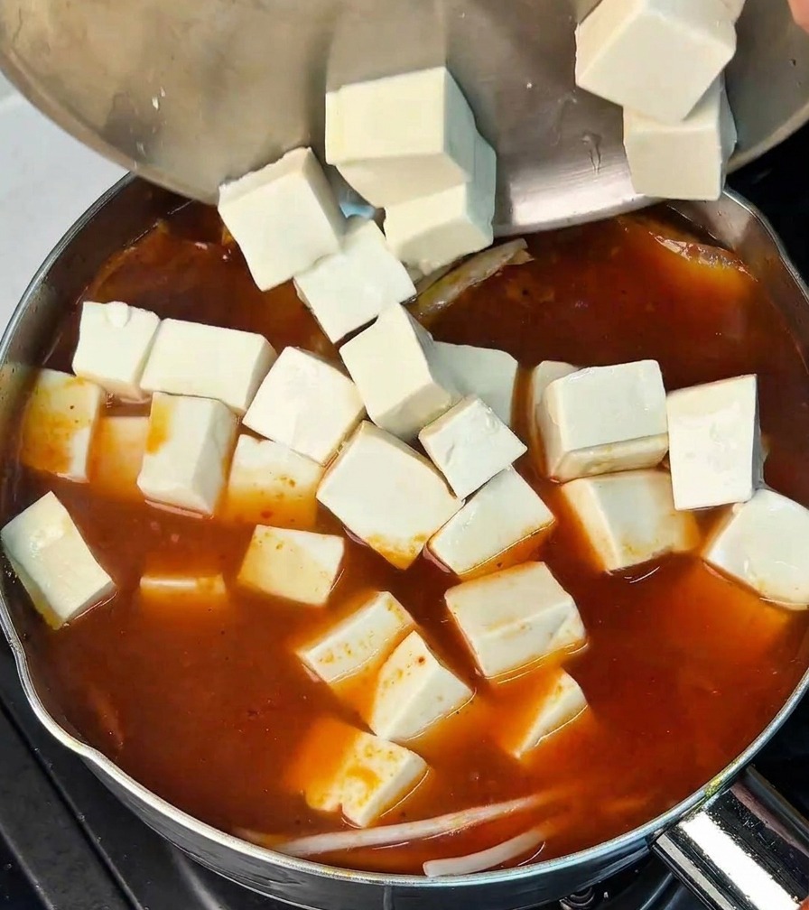 Numerous cubes of white tofu being poured from a stainless steel bowl into a pot of bubbling red kimchi broth.