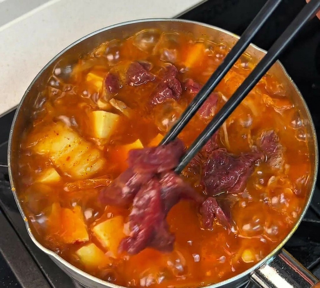 Slices of marinated raw beef being placed into a bubbling red soup with black chopsticks.