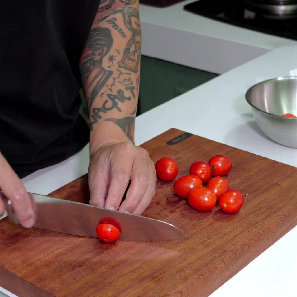 A chefs knife slicing through bright red cherry tomatoes on a dark wood cutting board.