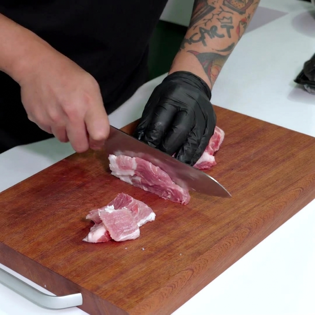 Hand in a black glove using a large knife to slice raw pink and white marbled pork belly into small strips on a wooden board.