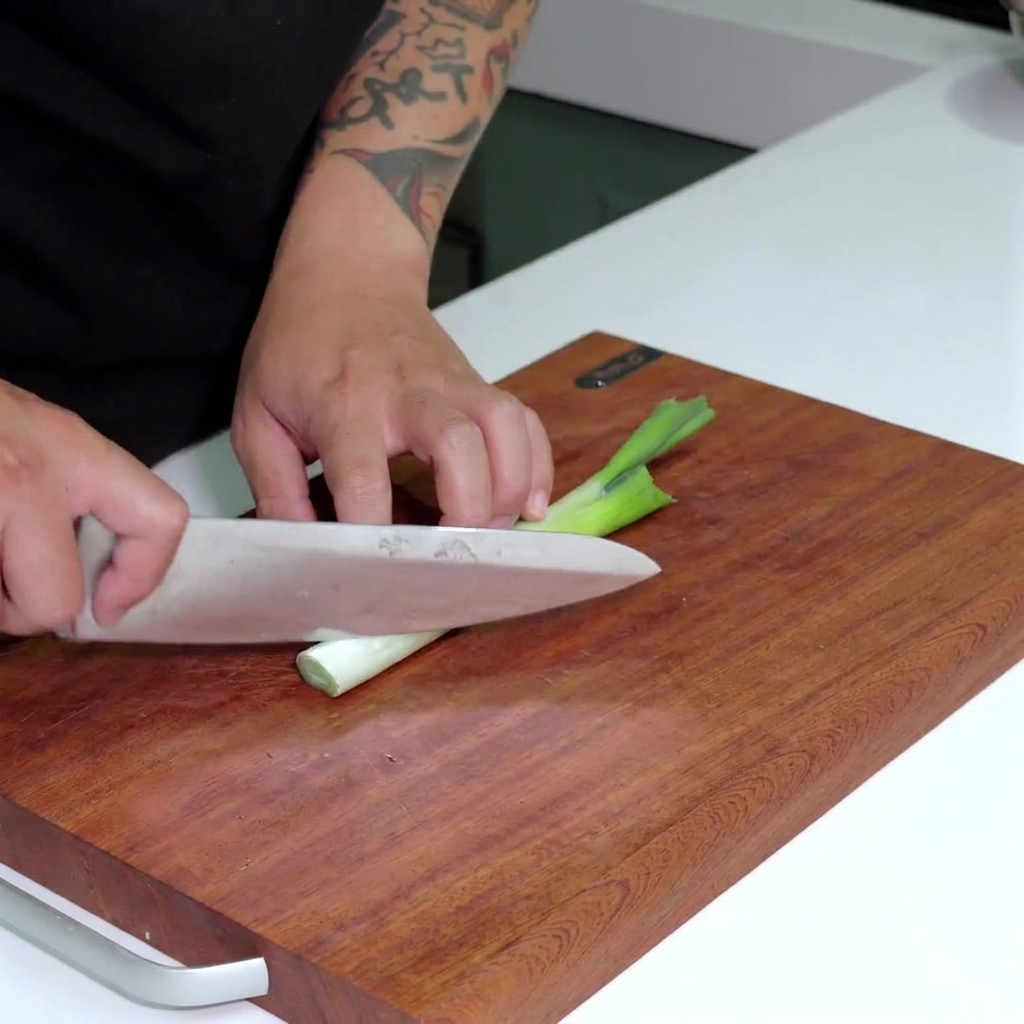 A knife slicing through a fresh white leek stalk into thin circular rounds on a wooden surface.