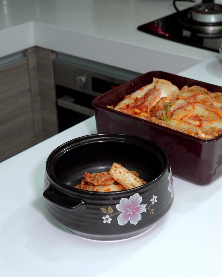 Fermented cabbage kimchi being cut with silver scissors inside a dark ceramic pot next to a container of kimchi.