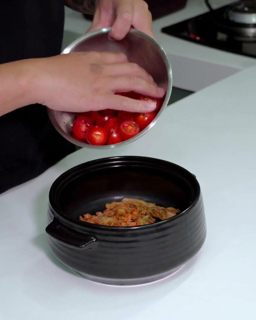 A person pouring a bowl of sliced red cherry tomatoes into a black pot already containing chopped kimchi.