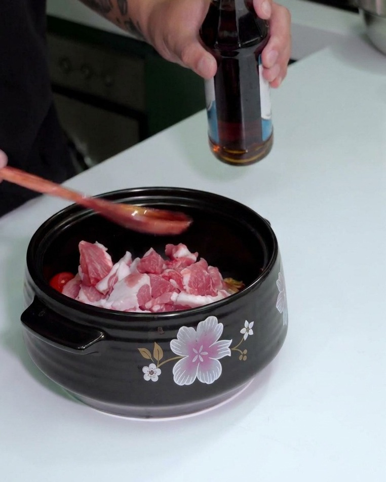 A person pouring fish sauce from a bottle into a wooden spoon held over a pot of ingredients.