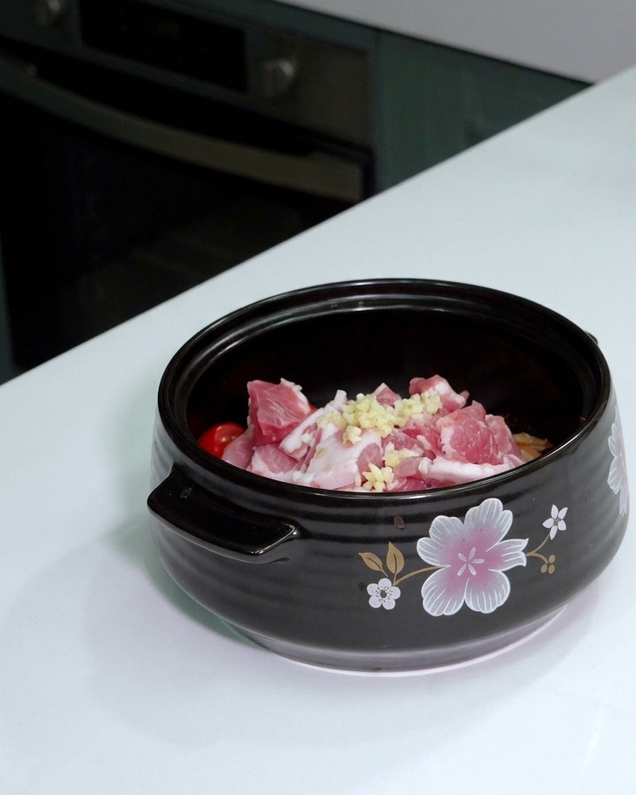 A spoonful of freshly minced garlic sitting on top of raw sliced pork belly inside a ceramic cooking pot.
